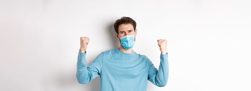 Covid-19, pandemic and social distancing concept. Happy young man in medical mask winning, screaming yes with satisfaction and raising hands up, celebrating victory, white background