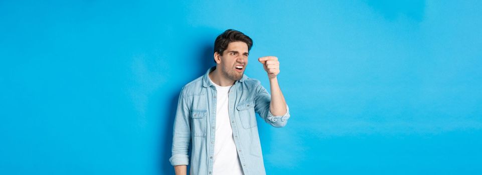 Confused guy holding something tiny in fingers and looking at it puzzled, standing over blue background