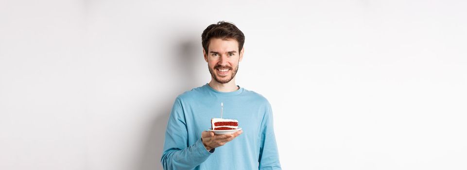 Celebration. Handsome young man celebrating birthday, holding bday cake with lit candle and smiling, making wish, standing over white background