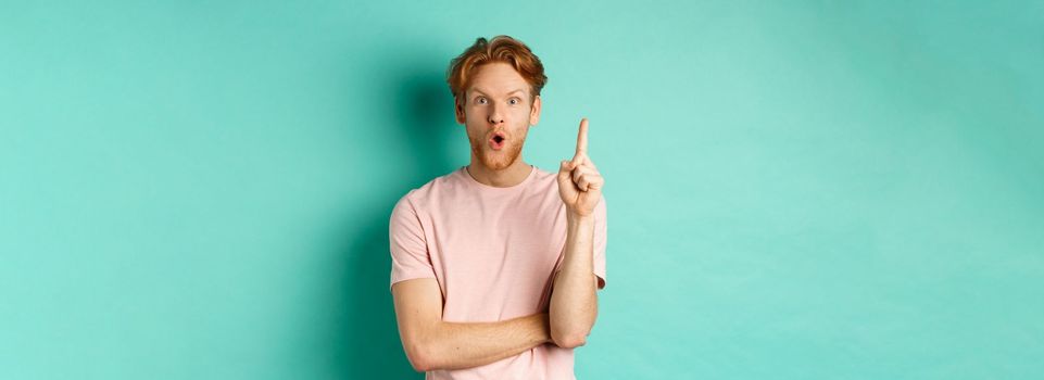 Pensive redhead man in t-shirt raising index finger, gasping as pitching at idea, saying suggestion, standing over turquoise background