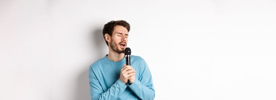 Young caucasian man singing song in microphone with carefree face, standing in karaoke over white background