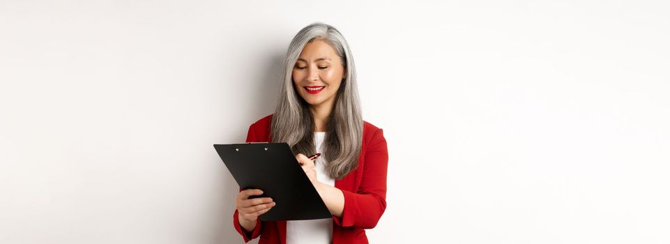 Successful asian senior businesswoman with grey hair taking notes on clipboard, inspecting enterprise, standing over white background