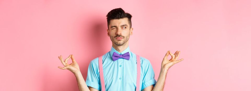 Funny guy with moustache and bow-tie fake meditating, peeking aside while doing yoga asana, standing over pink background