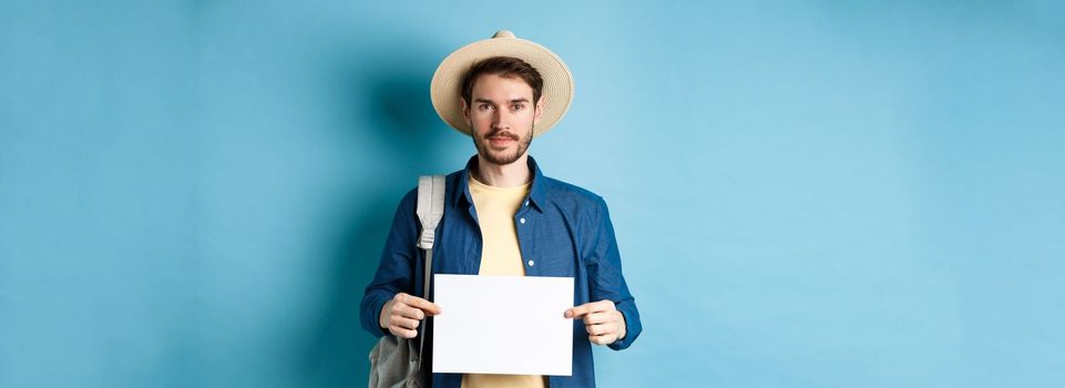 Image of smiling guy in straw hat backpacking, hitchhiking with piece of paper, travelling abroad on summer vacation, blue background