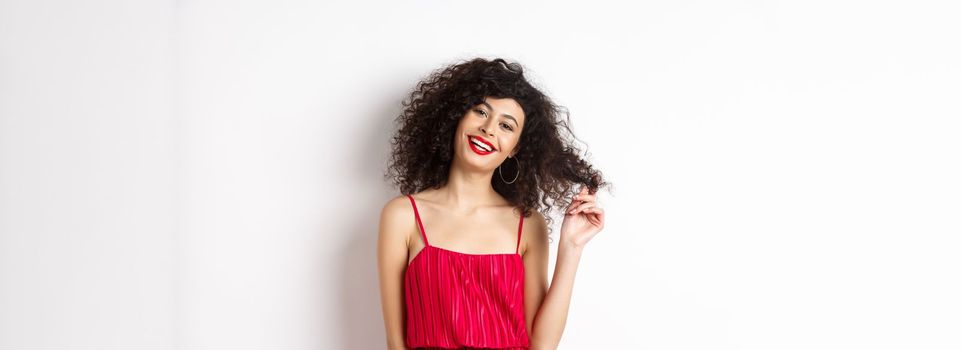 Beautiful woman with curly hair, wearing red dress and lipstick, playing with curl strand and smiling at camera, white background