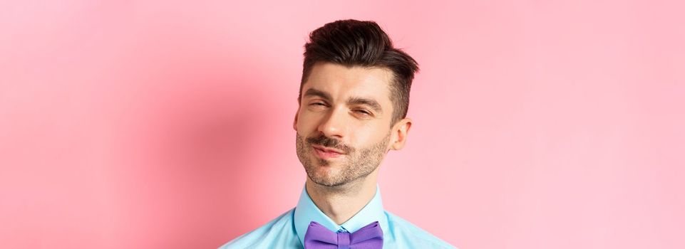 Close-up of smiling young guy with moustache, squinting intrigued, listening to something interesting, standing in bow-tie on pink background