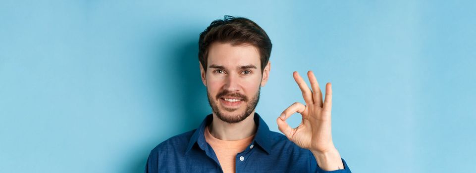 Close up of handsome bearded guy showing okay gesture and smiling, standing on blue background