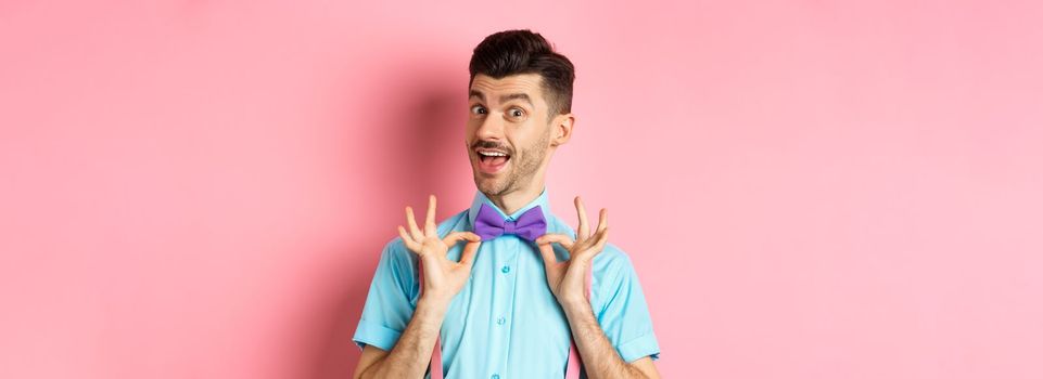 Classy handsome man fixing bow-tie on neck and smiling, getting ready for date or party, standing on pink background