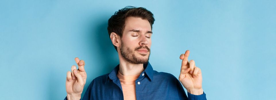 Close up of hopeful young man cross fingers for good luck and close eyes, making wish with calm expression, standing on blue background
