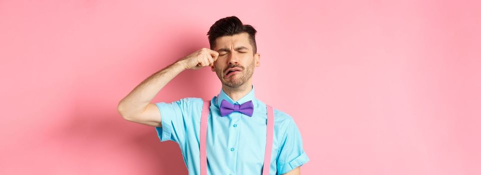 Image of heartbroken guy crying and wiping tear off face, sobbing and feeling sad or lonely, standing upset on pink background