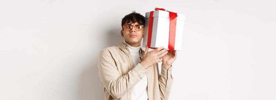Intrigued guy listening whats inside gift box, trying guess present, standing focused on white background.