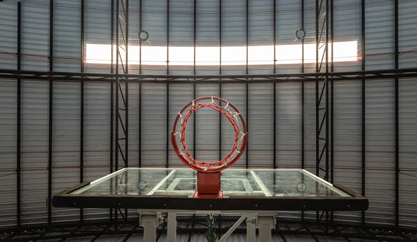 View from bottom of Basketball fiberglass backboard, Hoop red metal ring and white net against Ceiling sheet metal in the gym. 