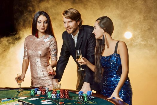 Two beautiful women and young man play on poker table in casino, focus on man and brunette