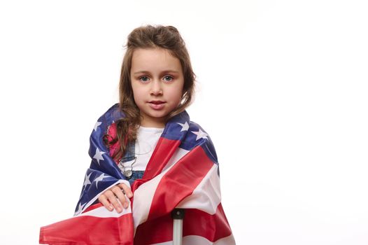 Happy preschooler girl wrapping in USA flag, smiling, posing with suitcase, isolated on white background, copy ad space