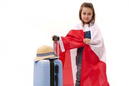 Cute little girl wrapping in Canada or Poland flag, smiles looking at camera, posing with suitcase over white background