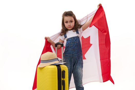 Little girl in white t-shirt and blue denim overalls, carrying Canada flag, poses with yellow suitcase, white background