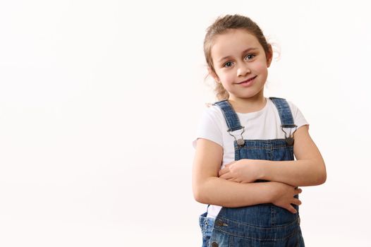 Cute baby girl in denim overalls, hugging herself, smiling toothy smile looking at camera, isolated on white background