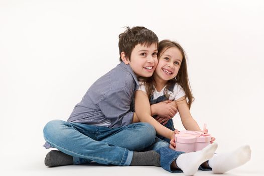 Cute kids wearing blue denim jeans, hold heart-shaped gift box, hugging each other, smiling at camera. White background