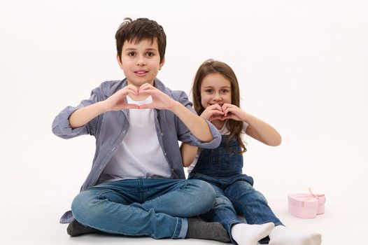 Adorable children, boy and girl showing a heart from fingers, smiling looking at camera, isolated over white background