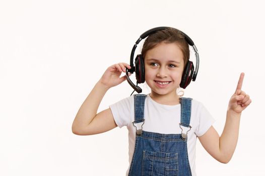 Smart little school girl, first grader wearing audio headset, cutely smiles looking at camera, pointing at a copy space on white background. Online meeting, education. Distance learning. Video call.