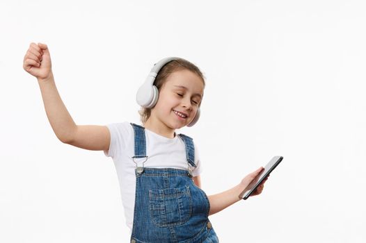 Cute baby girl in blue denim overall, holding smartphone and dancing while listening to the music on stereo headphones