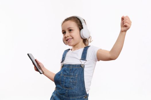 Joyful little girl with smartphone, listens music on her stereo headset, isolated over white background with copy space