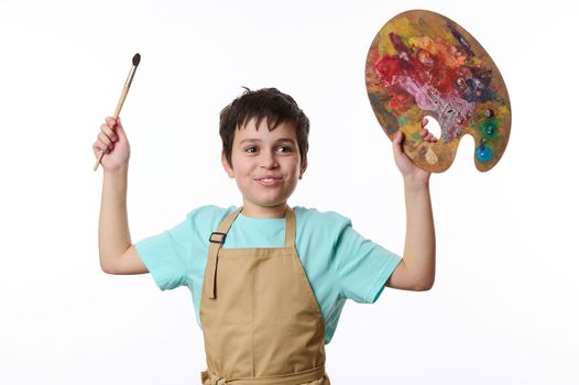 Inspired smiling teenage boy - talented painter in blue t-shirt and beige apron holding paintbrush and wooden palette