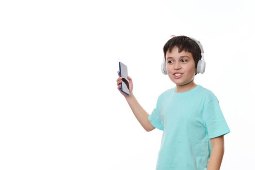 Teenage boy holds his smartphone and dances while listens to music on stereo headphones, isolated over white background