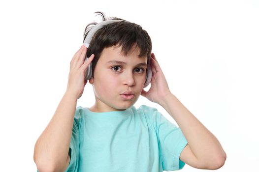 Close-up of a happy cute teenage boy puts on wireless stereo headphones, looking at camera, isolated on white background