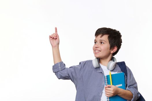 Cheerful teenage boy in casual wear, points at copy space on white background, posing with copybook and school backpack
