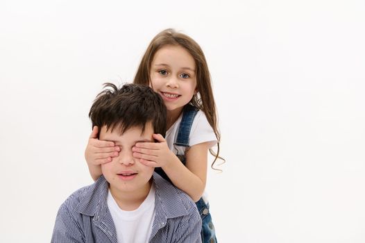 Adorable little child girl closing her brother's eyes with her hands, gently hugging him, isolated over white background