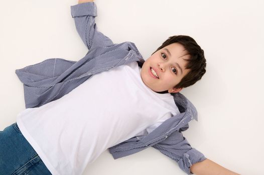 View from above of Caucasian adorable teen boy in blue jeans and white t-shirt, lying down on white background. Ad space