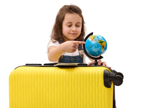 Happy European kid girl 5-6 years old with suitcase, pointing with her finger at the globe, isolated on white background