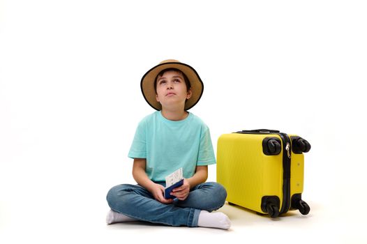 Multi-ethnic teenage traveler boy in straw hat with suitcase and boarding pass, dreamily looking at copy space