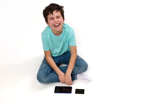 Top view of a dissatisfied Caucasian boy, expressing sadness sitting on white background with smartphone and credit card