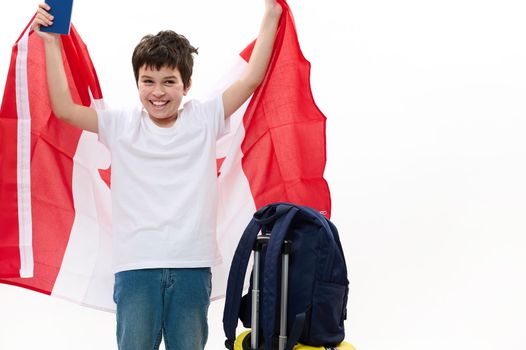Overjoyed American teenage boy with Canadian flag, boarding pass and luggage isolated over white background. Copy space