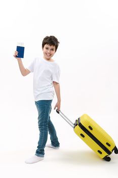 Full length portrait teenage boy in blue jeans and white t-shirt, with air ticket and suitcase going on summer vacations