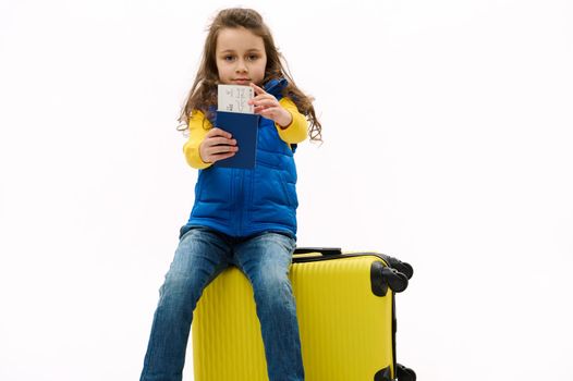 Cute kid girl holds out air ticket and boarding pass at camera, sitting on yellow travel bag, going for weekend getaway