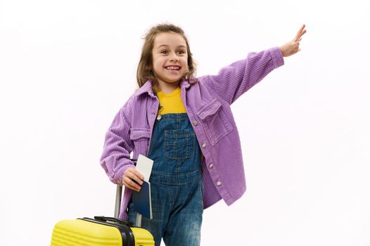Happy child girl in violet shirt with yellow suitcase, smiling, waving hello, isolated on white background. Copy space