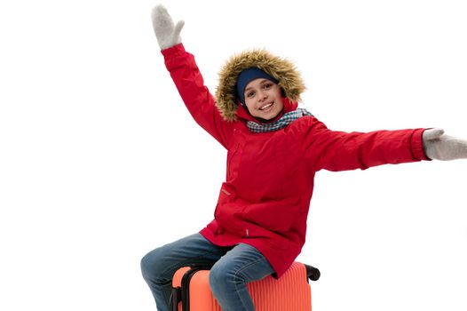 Smiling teenage traveler boy in warm winter clothes, sits on a suitcase, travels abroad, travelling on weekends getaway
