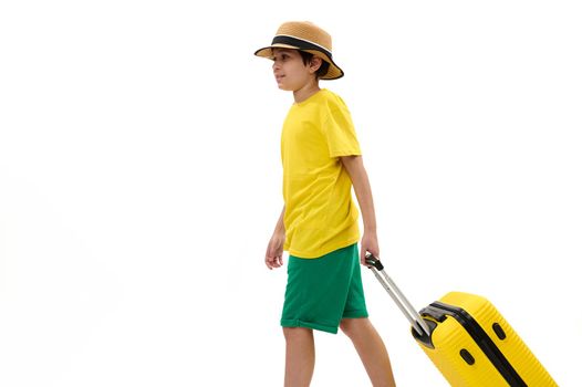 Teenager boy in yellow t-shirt and green shorts, walks with suitcase, traveling abroad for summer holidays. Copy space
