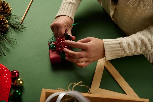 Close-up woman wrapping a small Christmas present in red gift paper and tying up with green decorative tape, tying a bow