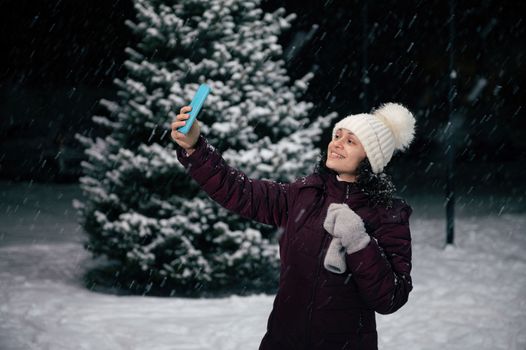 Multi-ethnic pretty woman in warm clothes smiles while taking selfie on her smartphone, enjoying a snowy winter evening