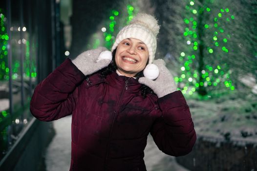 Delightful woman in warm clothes, smiling a beautiful toothy smile while playing with snowballs on snow covered street
