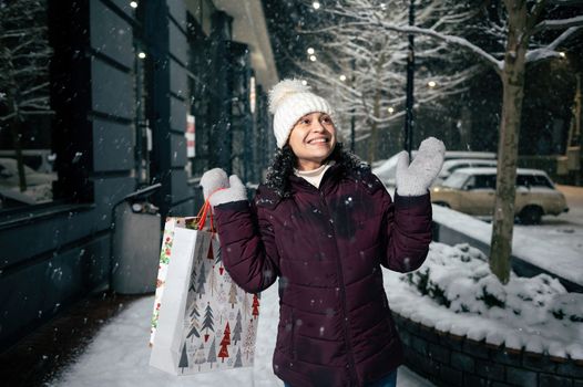 Delightful young woman catches snowflakes while walking down the snow covered street with shopping bags