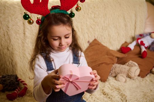 Charming Caucasian little girl wearing deer antler hoop, holding a pink heart shaped gift box. Boxing Day. Christmastime