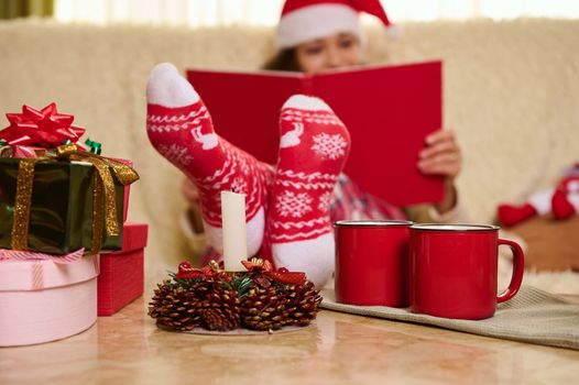 Focus on red socks with Christmas pattern, on blurred background of woman in Santa hat reading book at winter holidays
