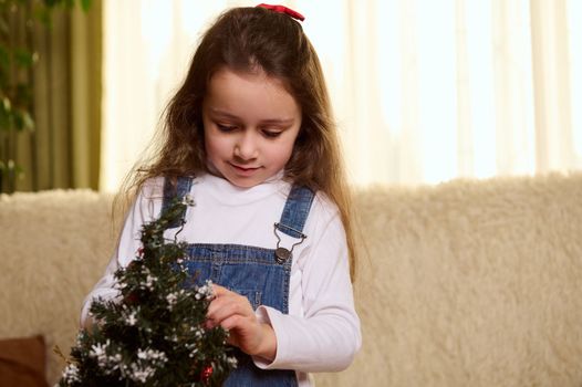 Close-up portrait of pretty child, beautiful little girl decorating a Christmas tree in the home interior. Christmastime