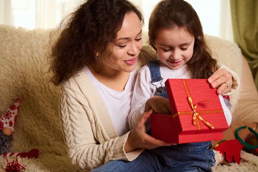 Multi-ethnic pretty woman, loving mother hugging her adorable lovely daughter while giving her a cute Christmas present.