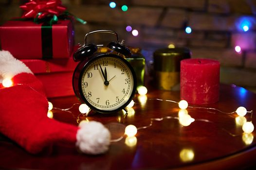 Midnight on alarm clock's dial, lit garlands, candle and gifts. Magic New Year atmosphere. Christmas